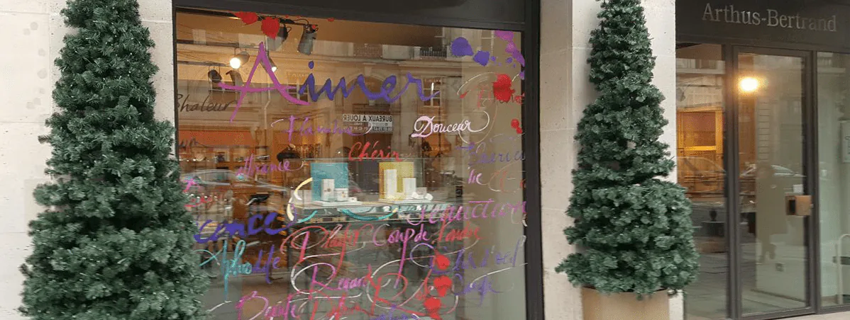 Vitrophanie de fin d'année Arthus-Bertrand : mots calligraphiés sur la vitrine entre deux sapins de Noël