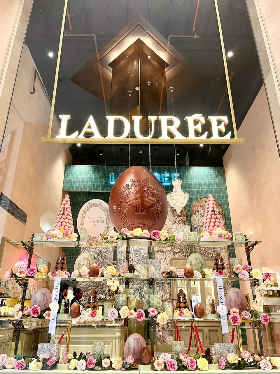 Vitrine de Pâques Ladurée sous l’enseigne lumineuse : œuf géant en chocolat, pyramides de macarons et roses