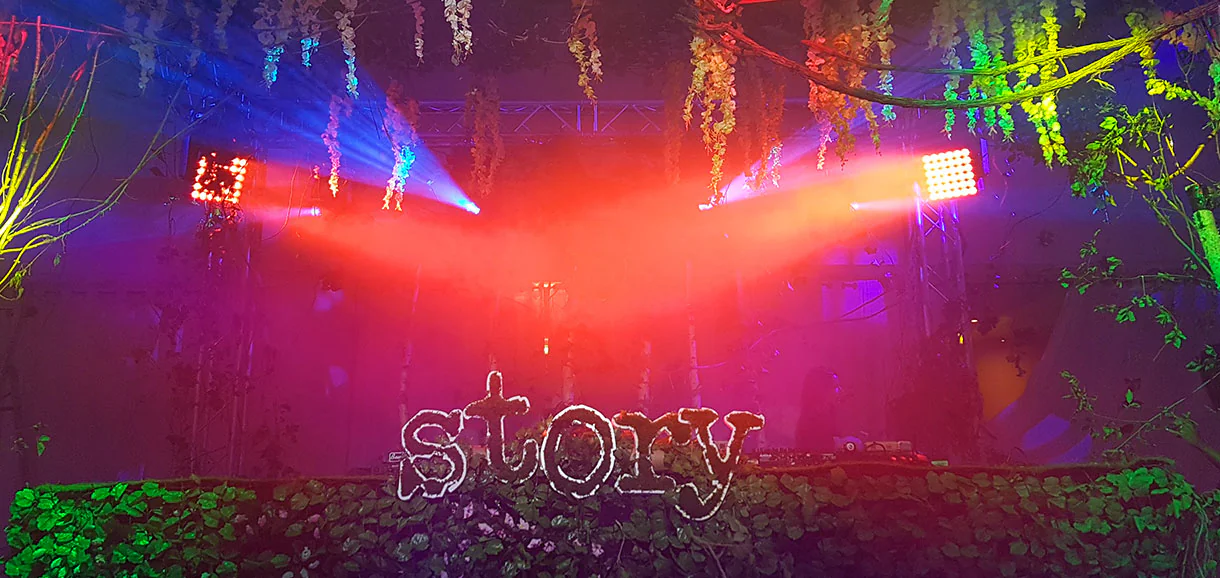 Logo Story en lettres lumineuses sur une haie de végétal stabilisé, sous projecteurs rouges et lianes suspendues