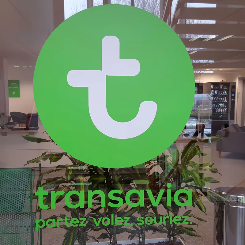 Logo Transavia et slogan « partez, volez, souriez » en vitrophanie verte sur une paroi vitrée de la cafétéria